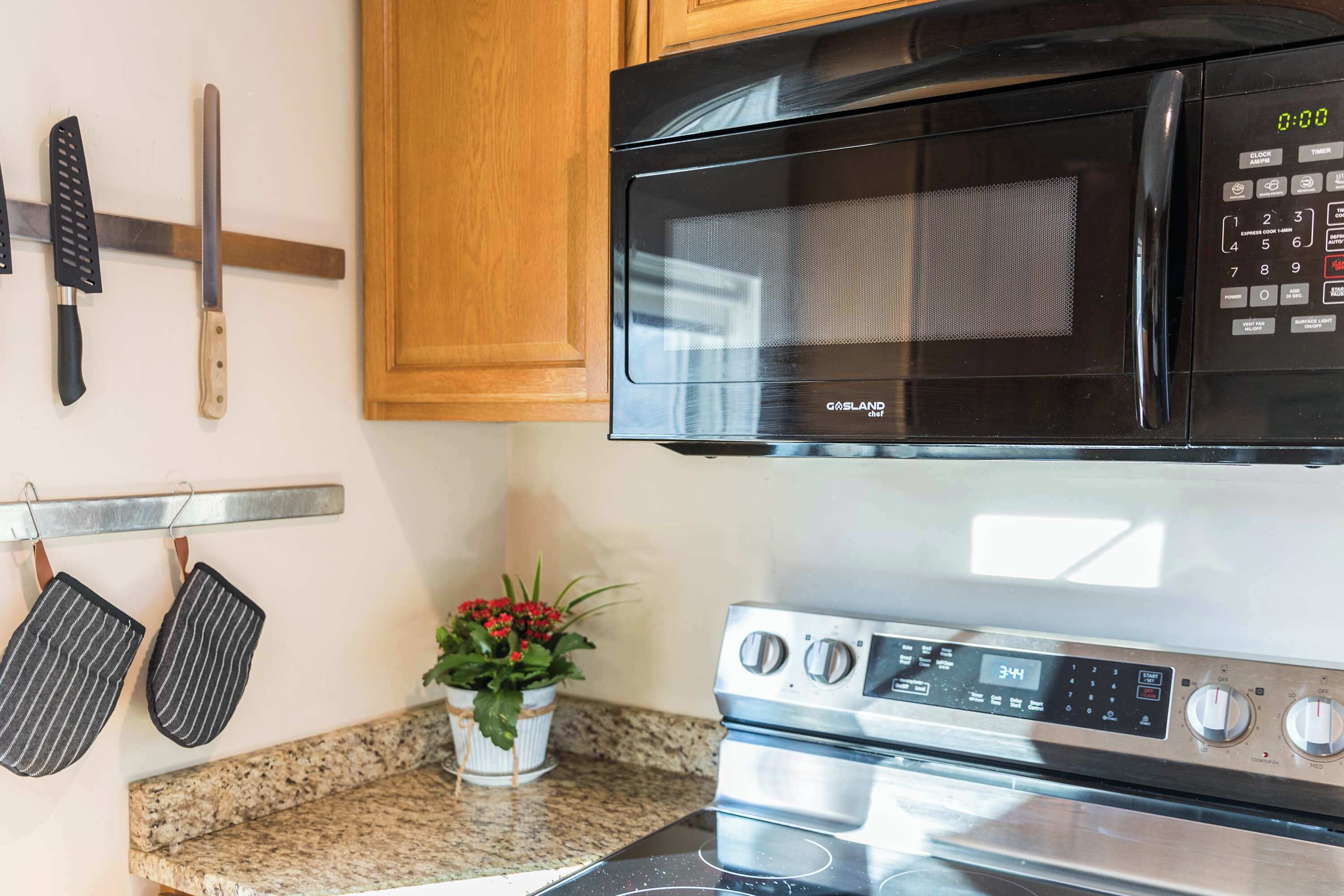 A kitchen with a black microwave, stainless steel stove, hanging knives, and striped oven mitts.