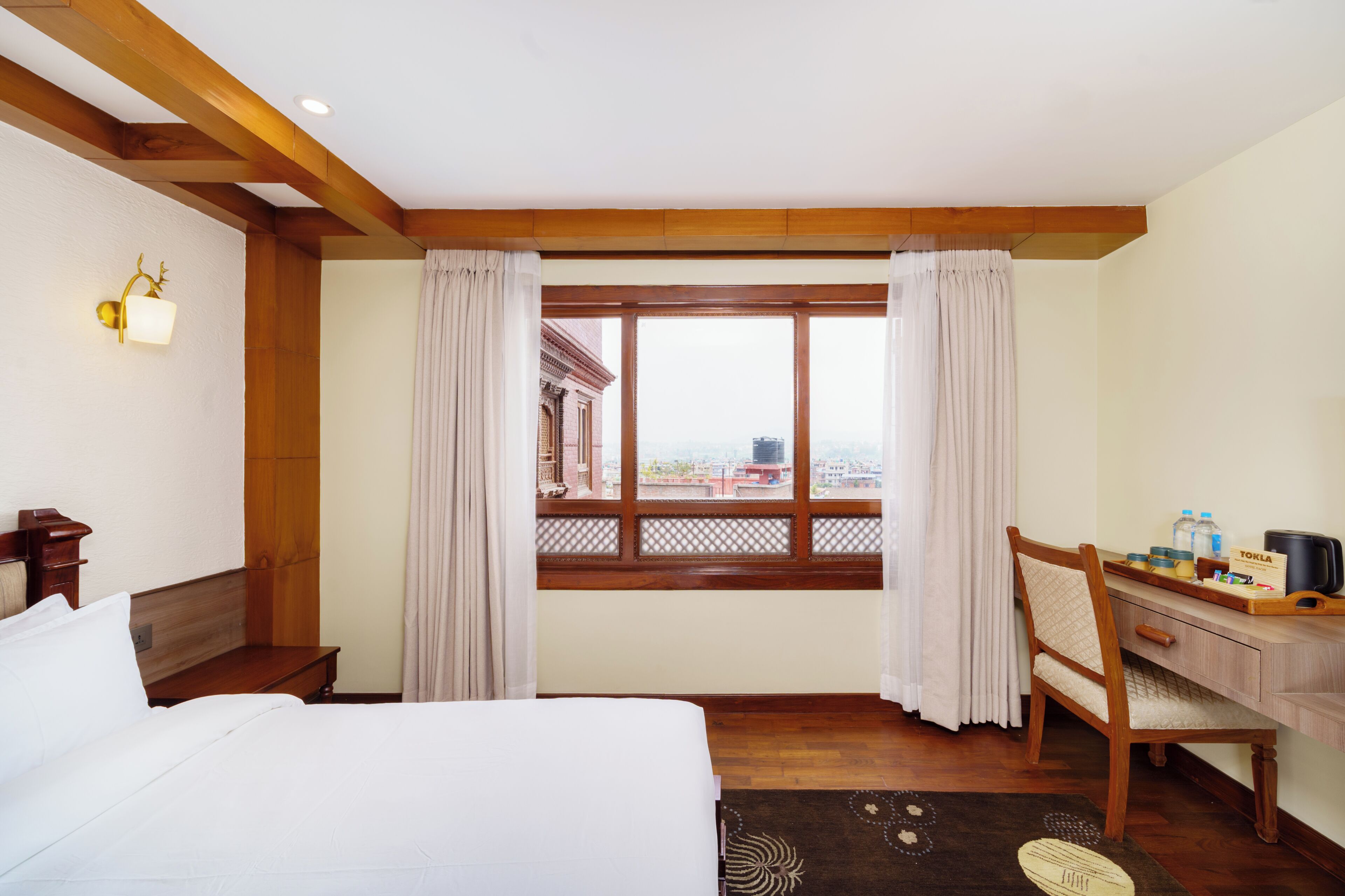 Ein Hotelzimmer mit Bett, Schreibtisch, Stuhl, einem Fenster mit Vorhängen und Blick auf Gebäude.