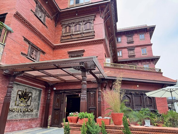 Un bâtiment traditionnel orné de sculptures complexes en bois, une enseigne indiquant « HÔTEL DACHI », et des plantes en pot devant l’entrée.