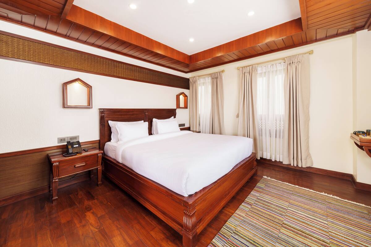 A hotel room with a large bed, wooden headboard, a nightstand with a phone, and a window with curtains.