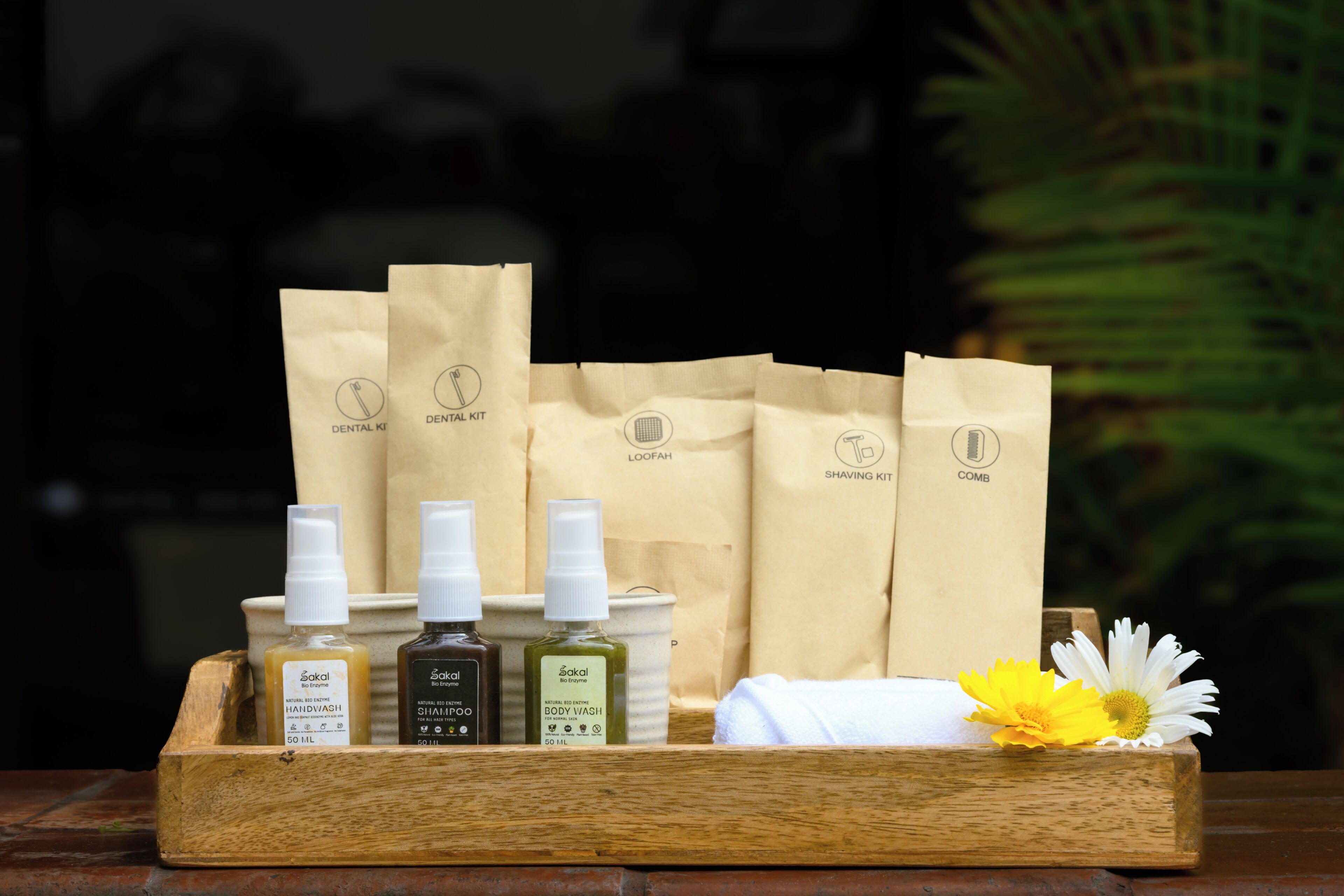 A wooden tray with three bottles of handwash, a bottle of body wash, and a shaving kit, along with a white towel and a daisy.