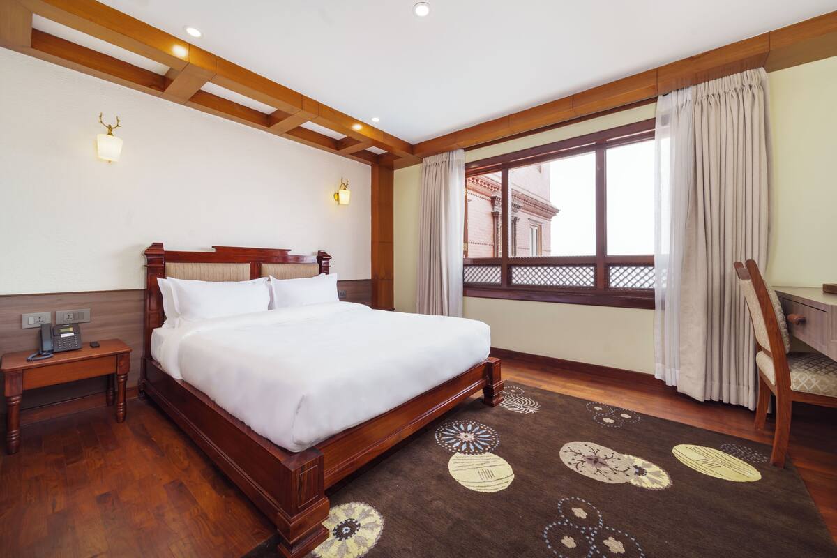 A hotel room with a wooden bed, a nightstand, a desk, a chair, a window with curtains, and a patterned rug.