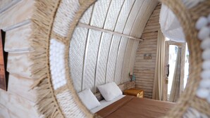 Um espelho circular reflete uma cama com almofadas brancas, uma cabeceira de madeira e uma mesa de cabeceira de madeira com uma luminária.