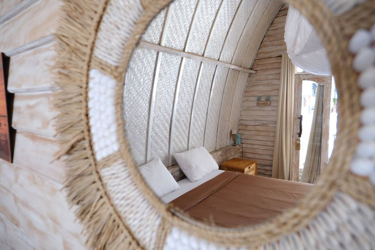 Een ronde spiegel toont een bed met witte kussens, een houten hoofdbord en een nachtkastje met een lamp.