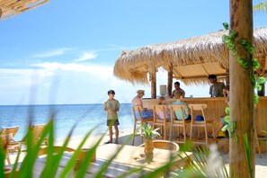 Un bar frente a la playa con sillas de madera y techo de paja, con vista al océano.