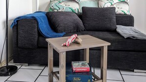 Een woonkamer met een donkergrijze bank, gekleurde kussens, een klein houten bijzettafeltje en een Monopoly spel op tafel.