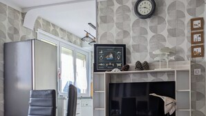 Een moderne woonkamer met een eethoek, met een televisie, planken met decoratieve objecten en een klok aan de muur.