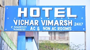 Un letrero azul de hotel con el nombre 'VICHAR VIMARSH' y la inscripción '24X7 Habitaciones con aire acondicionado y sin aire acondicionado', indicando la disponibilidad de ambos tipos de habitaciones.