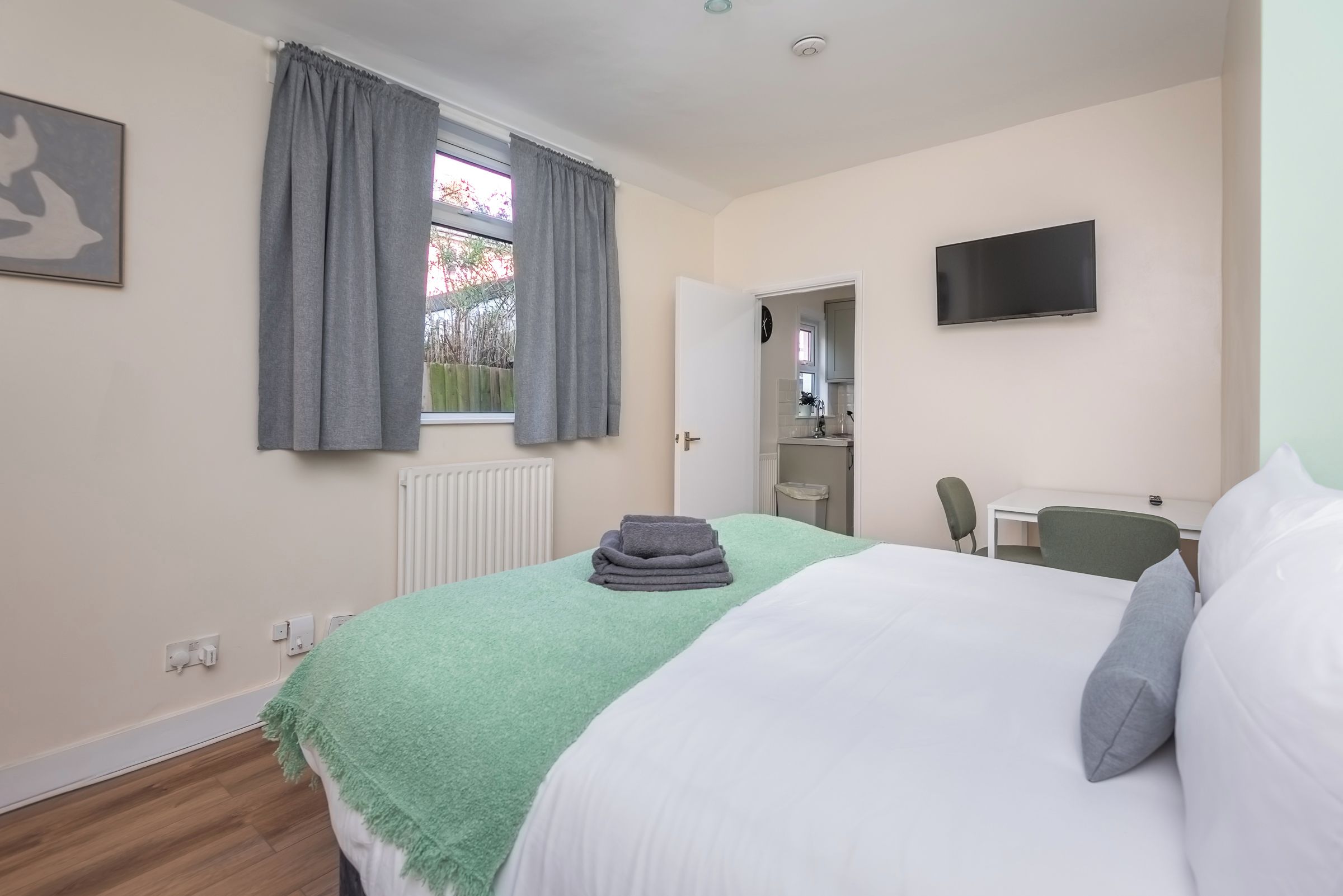 Ein Schlafzimmer mit Bett, Fernseher, Fenster mit Vorhängen, Heizkörper und einer Tür zu einem Badezimmer.