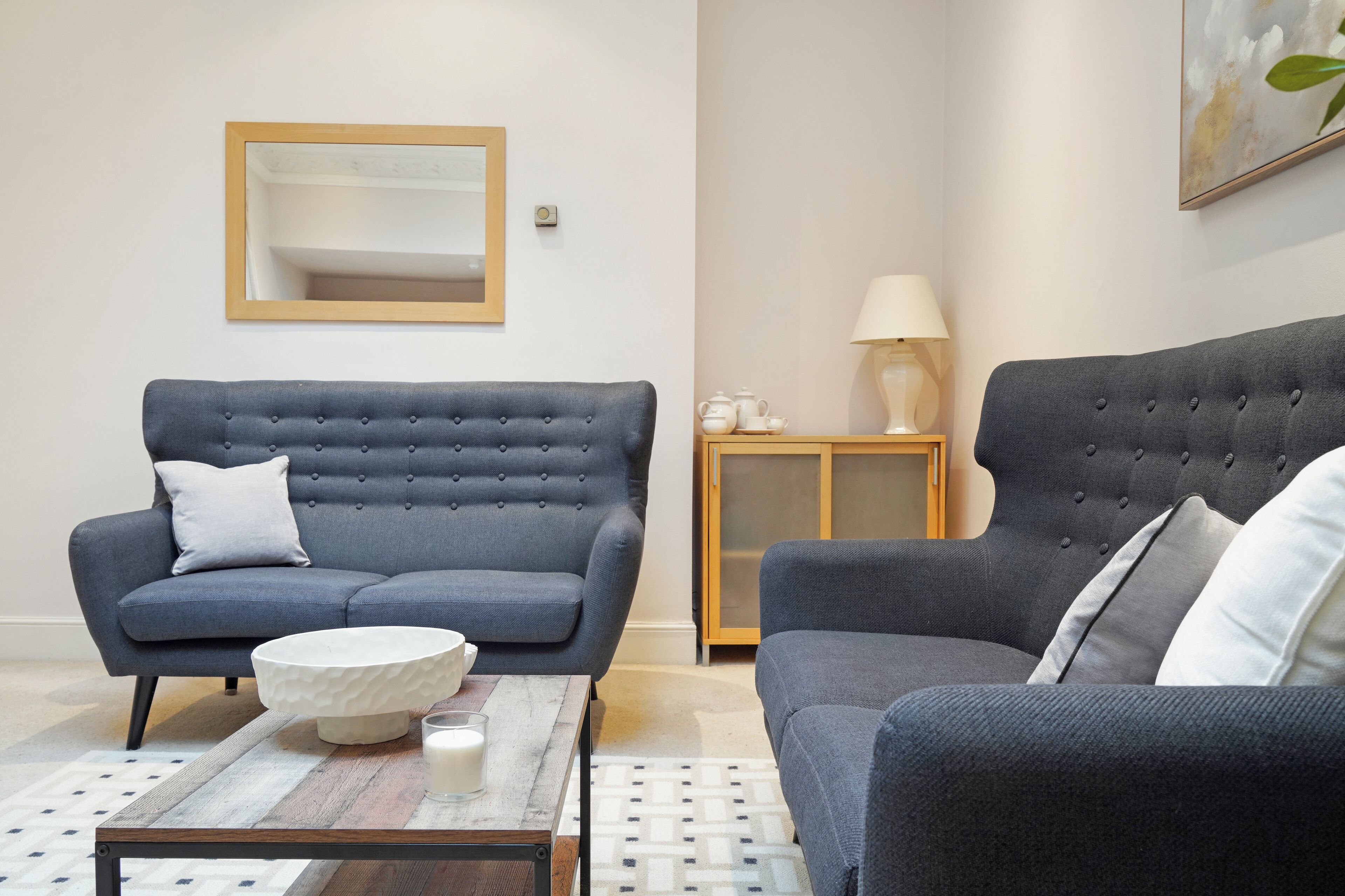 A modern living room with two dark grey sofas, a wooden coffee table with a white bowl and glass, and a small cabinet with a lamp.