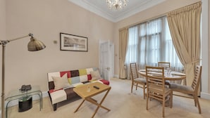 A living room with a sofa, a coffee table, a dining table with chairs, a chandelier, and a window with curtains. - Hertford Flat 4 (London)