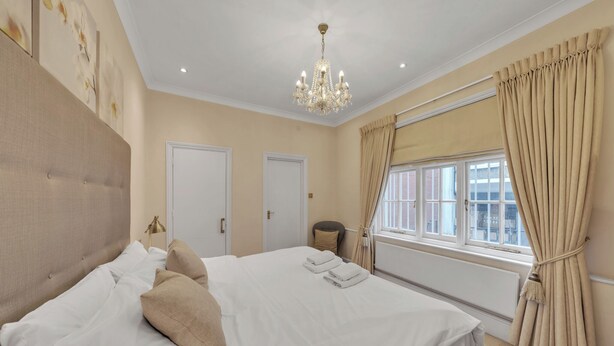 A well-lit bedroom with a large bed, a chandelier, beige curtains, and a view of the city through the window. - Hertford Flat 4 (London)