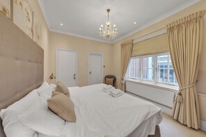 A well-lit bedroom with a large bed, a chandelier, beige curtains, and a view of the city through the window. - Hertford Flat 4 (London)