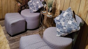 Ein gemütlicher Sitzbereich mit gepolsterten Hocker und Sesseln, einem flachbildfernseher an der Wand und einem dekorativen Regal mit Büchern und einer Schale.