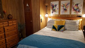 Ein Schlafzimmer mit einem hölzernen Kopfteil, einem Bett mit blau-weißer Streifenauflage und drei an der Wand befestigten Bildern von Kolibris.