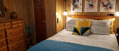 A bedroom with a wooden headboard, a bed with a blue and white striped bedspread, and three wall-mounted paintings of hummingbirds.