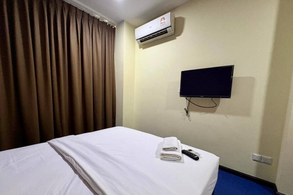 A hotel room with a bed, an air conditioner, a TV, and a remote control on the bed.