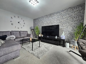 A modern living room with a grey sofa, a coffee table, a flat-screen TV, a stone wall, and a lamp.