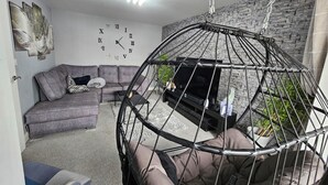 Ein Wohnzimmer mit grauem Sofa, einem schwarzen Fernseher, einem Hängesessel und einer Wanduhr.