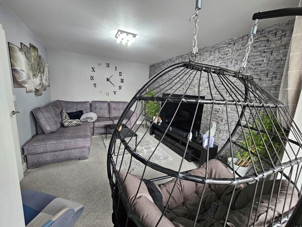 A living room with a grey sofa, a black TV, a hanging chair, and a wall clock.