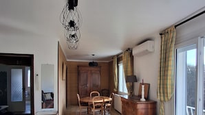 Ein geräumiger Wohnbereich mit Esstisch, Stühlen, einem schwarzen Sofa, einem kleinen Tisch mit Lampe und einem großen Fenster mit Vorhängen.