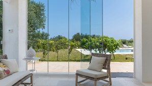 A room with a large sliding glass door leading to a pool and garden. There is a wicker chair and a small table with a decorative item. - Exclusive, Modern Country House with Private Pool – Your Dream Holiday in Puglia (Puglia)