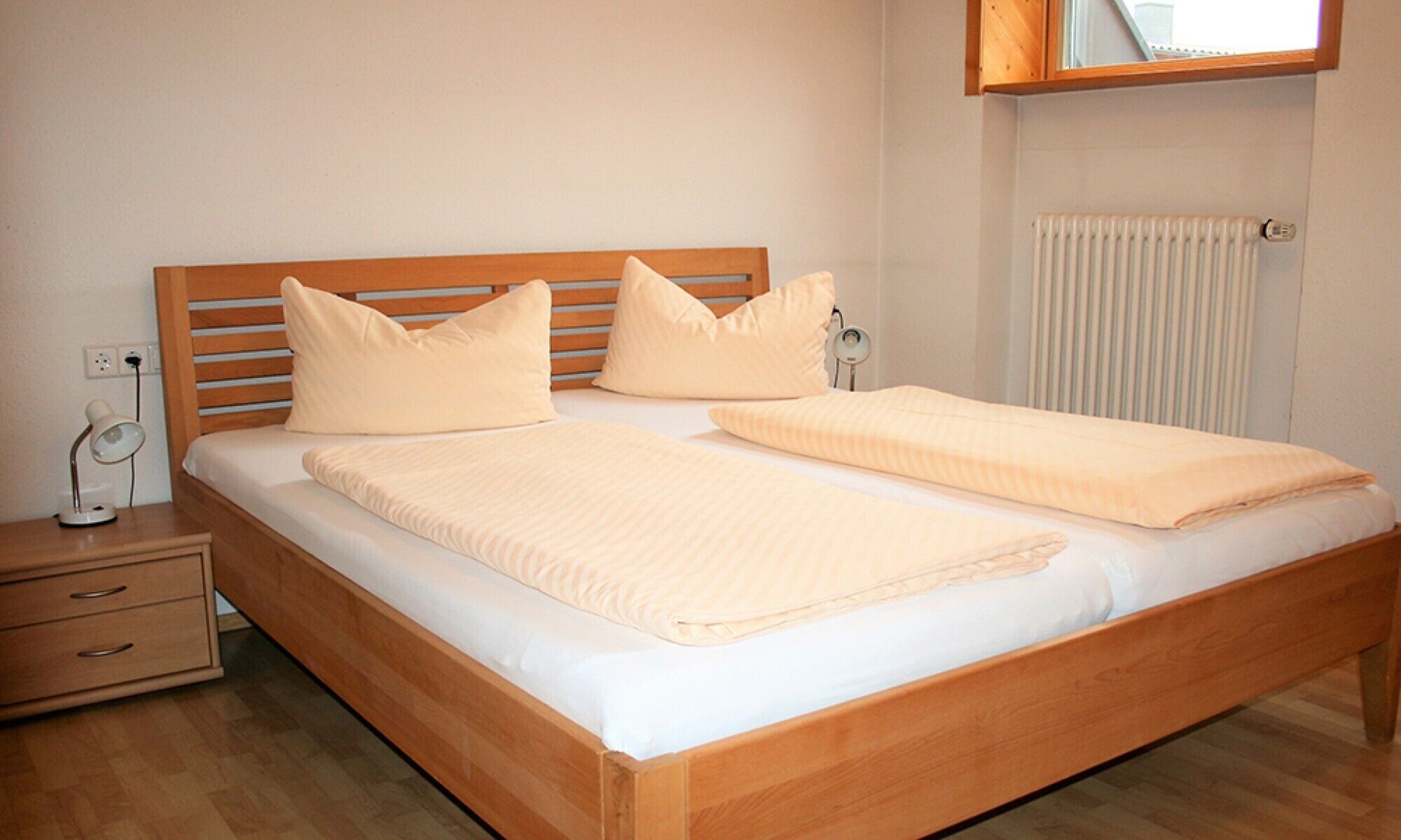 Ein Doppelbett mit hölzernem Kopfteil, ein Nachttisch mit Lampe und ein Heizkörper an der Wand.