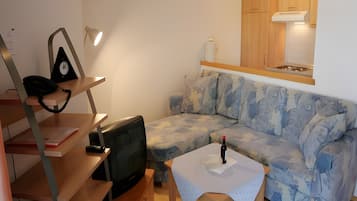 Ein kleiner, möblierter Raum mit einer Couch, einem Fernseher, einem Tisch mit einer Flasche und Holzschränken über der Couch.