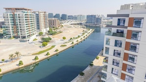 A modern high-rise building with a waterfront view, featuring a promenade along the water, a bridge in the background, and other high-rise buildings. - Bloomfields 1BR Canal The View Raha (Abu Dhabi)