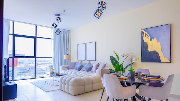 A modern living room with a large sofa, dining table, and chairs. There is a large window with a view of the city. - Bloomfields 1BR Canal The View Raha (Abu Dhabi)