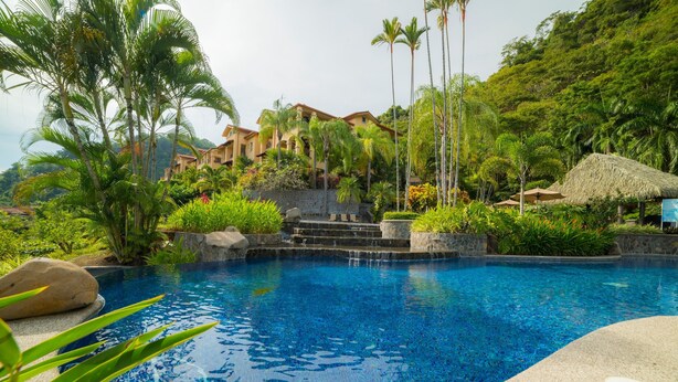 A swimming pool with clear blue water, surrounded by lush greenery and palm trees. - Marbella 1E 3 bdr Ocean View in Los Suenos (Jacó)