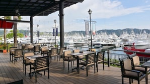 Zona de comedor al aire libre con mesas y sillas, con vistas a un puerto deportivo con numerosos barcos.