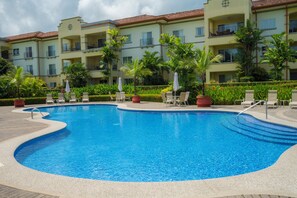 Ein Pool mit Liegestühlen und Sonnenschirmen, umgeben von einem angelegten Garten und einem mehrstöckigen Gebäude mit Balkonen.