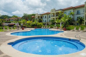 Ein Pool mit Liegestühlen und Sonnenschirmen, umgeben von einem angelegten Garten und Hotelgebäuden.