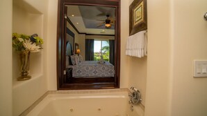 Ein Badezimmer mit einer großen Badewanne, einem Deckenventilator, einem gerahmten Bild an der Wand und Blick ins Freie.