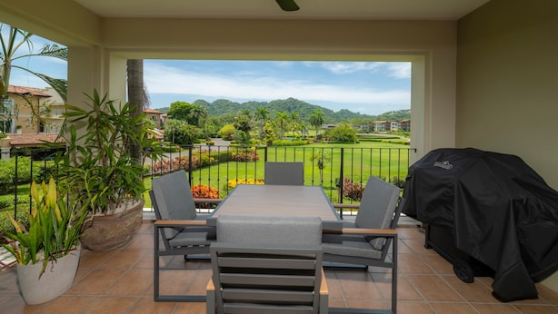 A balcony with a table and chairs, a ceiling fan, and a view of a landscaped garden and distant hills. - Del Mar 5J 3 bdr Garden View in Los Suenos (Jacó)