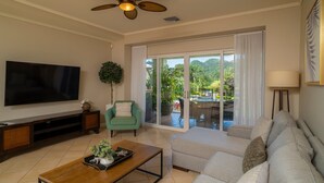 A living room with a flat-screen TV, a ceiling fan, a sofa, a coffee table, and a sliding glass door leading to a balcony with a view of greenery. - Del Mar 5J 3 bdr Garden View in Los Suenos (Jacó)