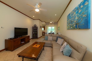 A living room with a TV, wooden entertainment unit, beige sofa, and a painting on the wall. - Veranda 8H 3 bdr Garden View in Los Suenos (Jacó)