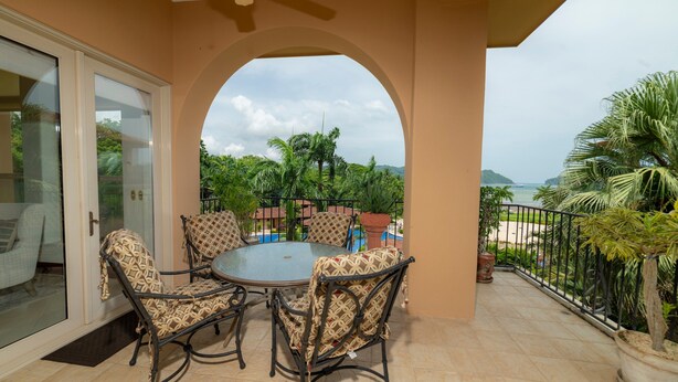 A covered patio with a glass door leading to a pool area, surrounded by palm trees and a view of the ocean. - Bella Vista 3A 3 Bdrs Ocean View in Los Suenos (Jacó)