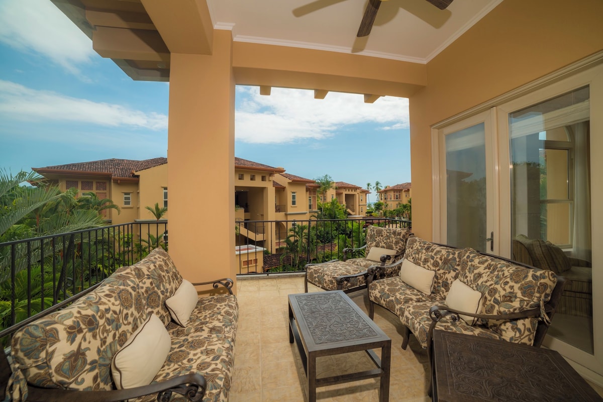 A balcony with a sofa set and a small table, overlooking a residential area with similar buildings.