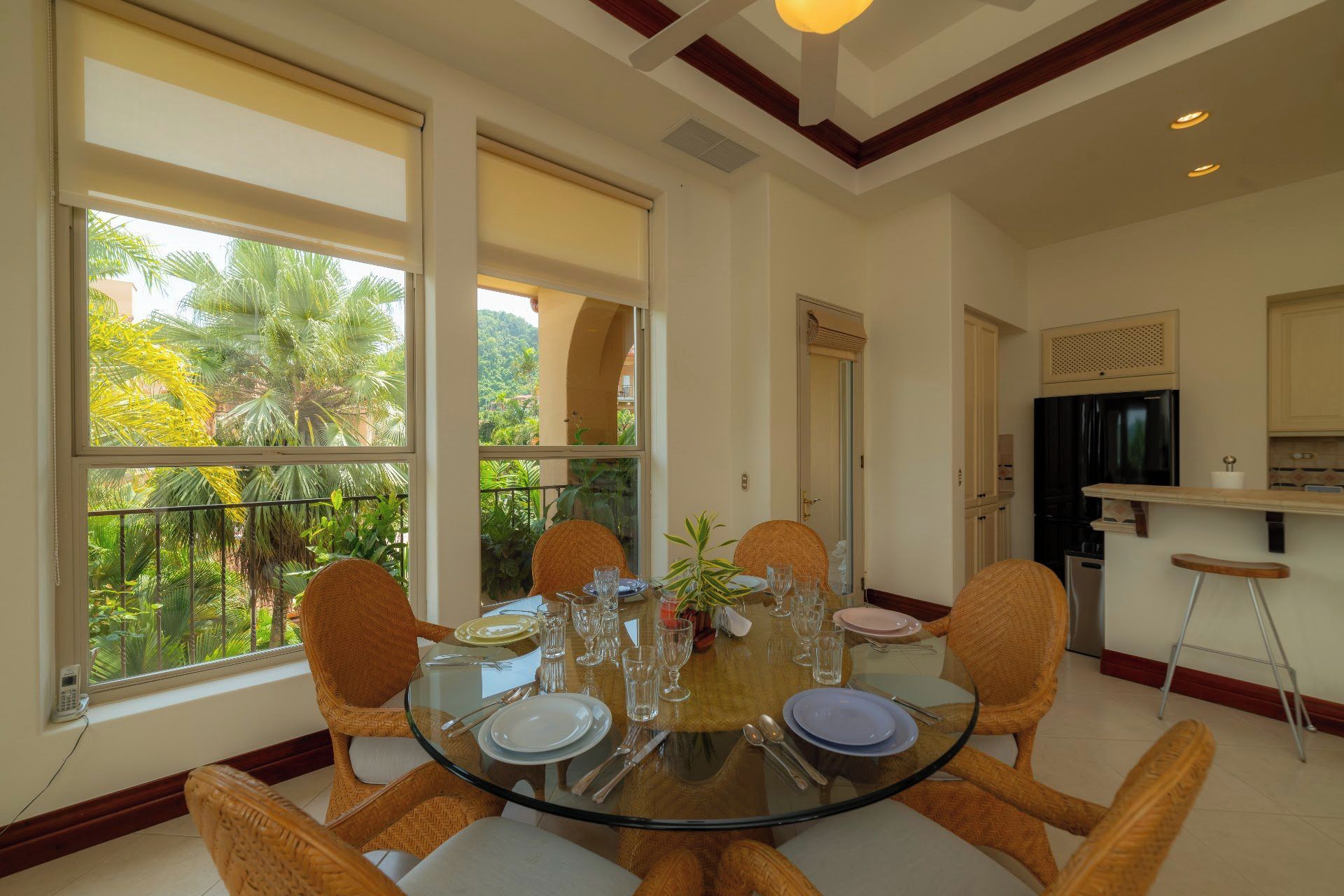 A dining area with a round glass table, six wicker chairs, and a view of greenery through large windows.