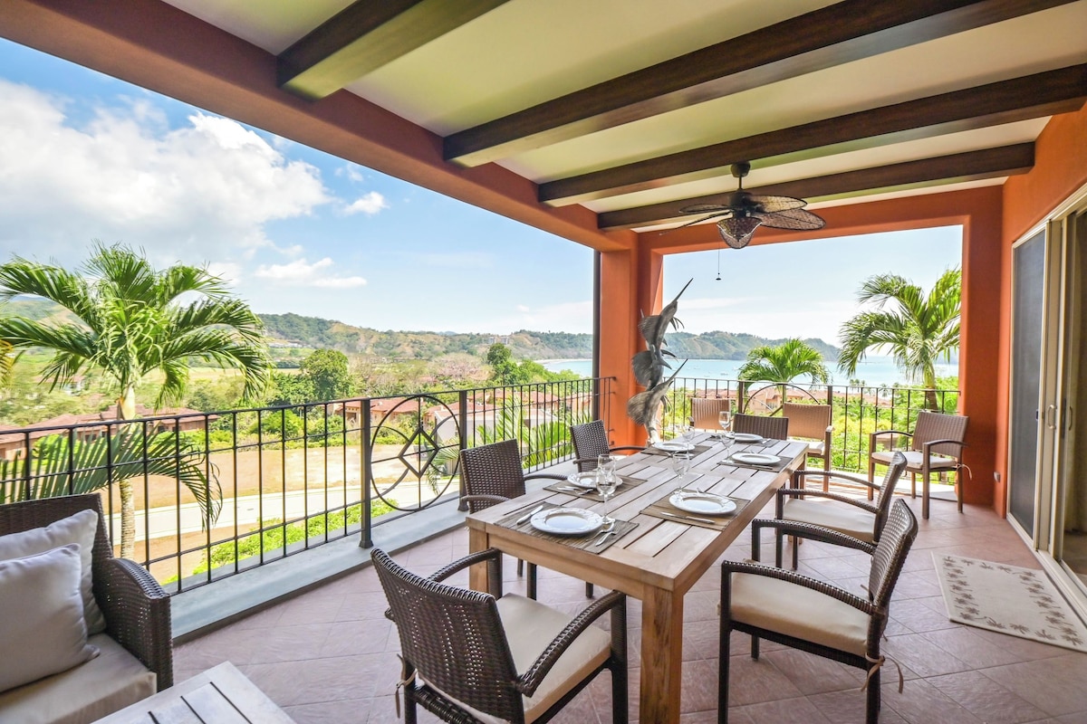 A covered outdoor dining area with a table set for two, overlooking a scenic landscape with hills and palm trees.
