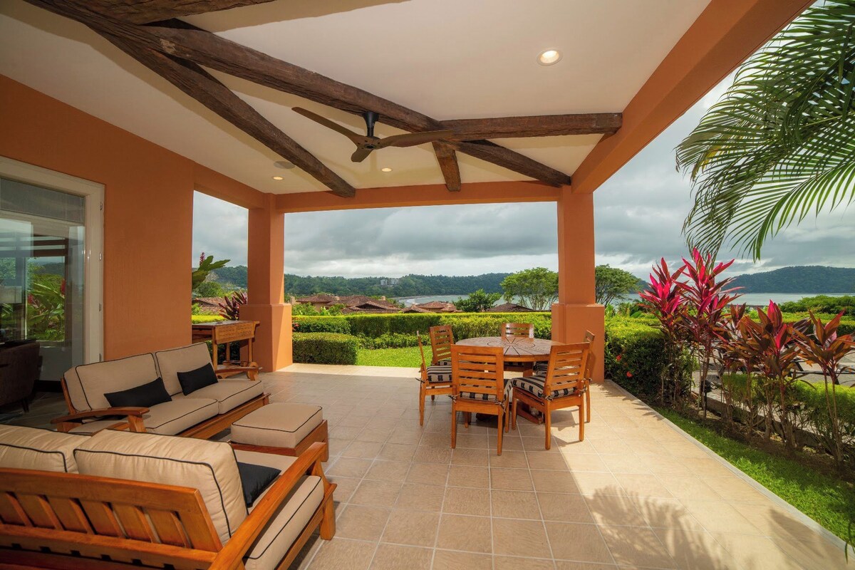A covered outdoor seating area with a table and chairs, a sofa, and a view of a landscape with greenery and a body of water.