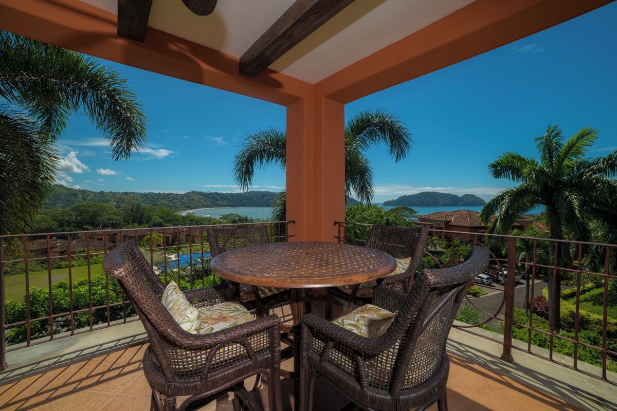 A balcony with a round table and wicker chairs, overlooking a scenic view of a lake and mountains.