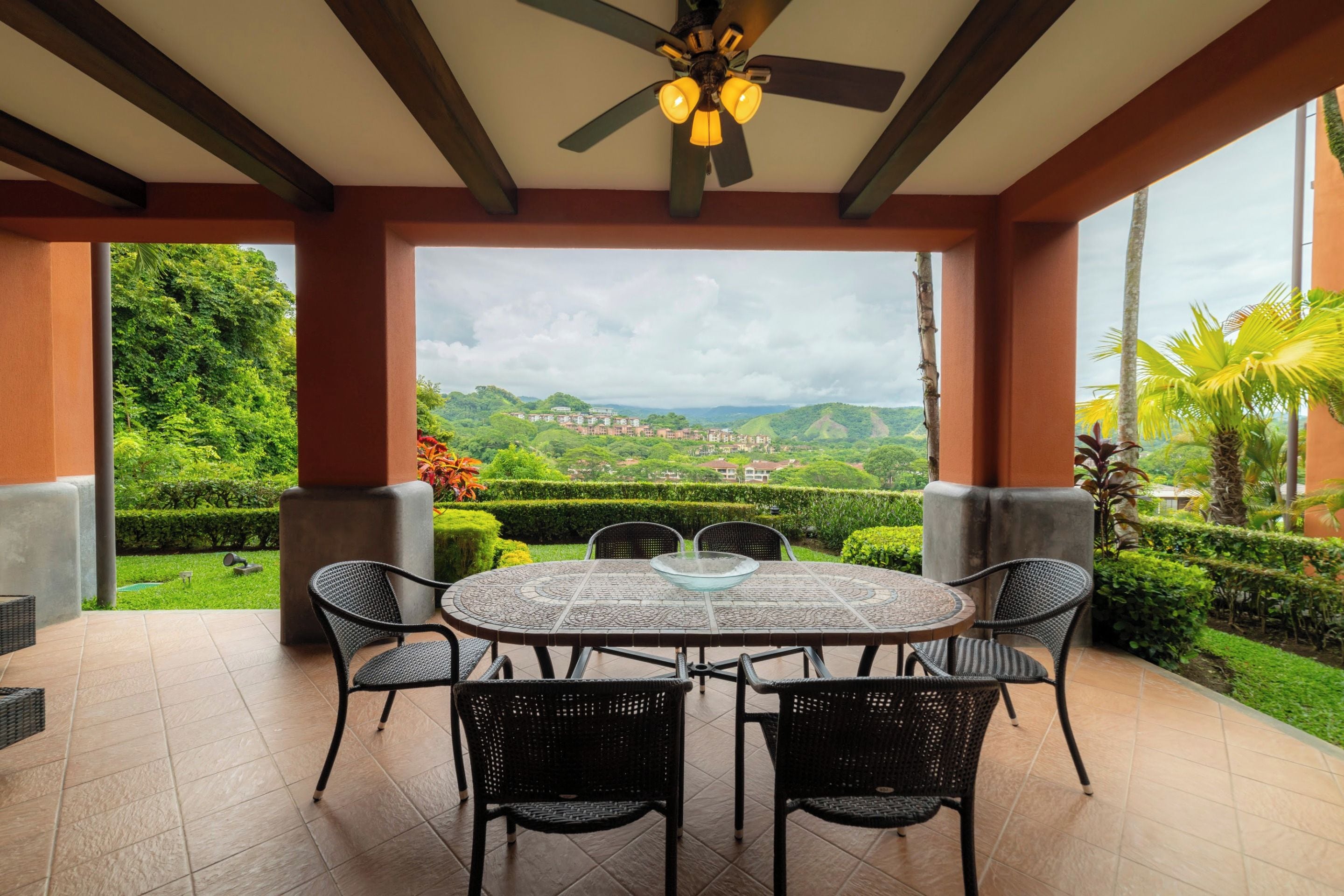 Uma área de estar coberta ao ar livre com mesa e cadeiras, ventilador de teto e vista para uma paisagem exuberante com edifícios ao fundo.