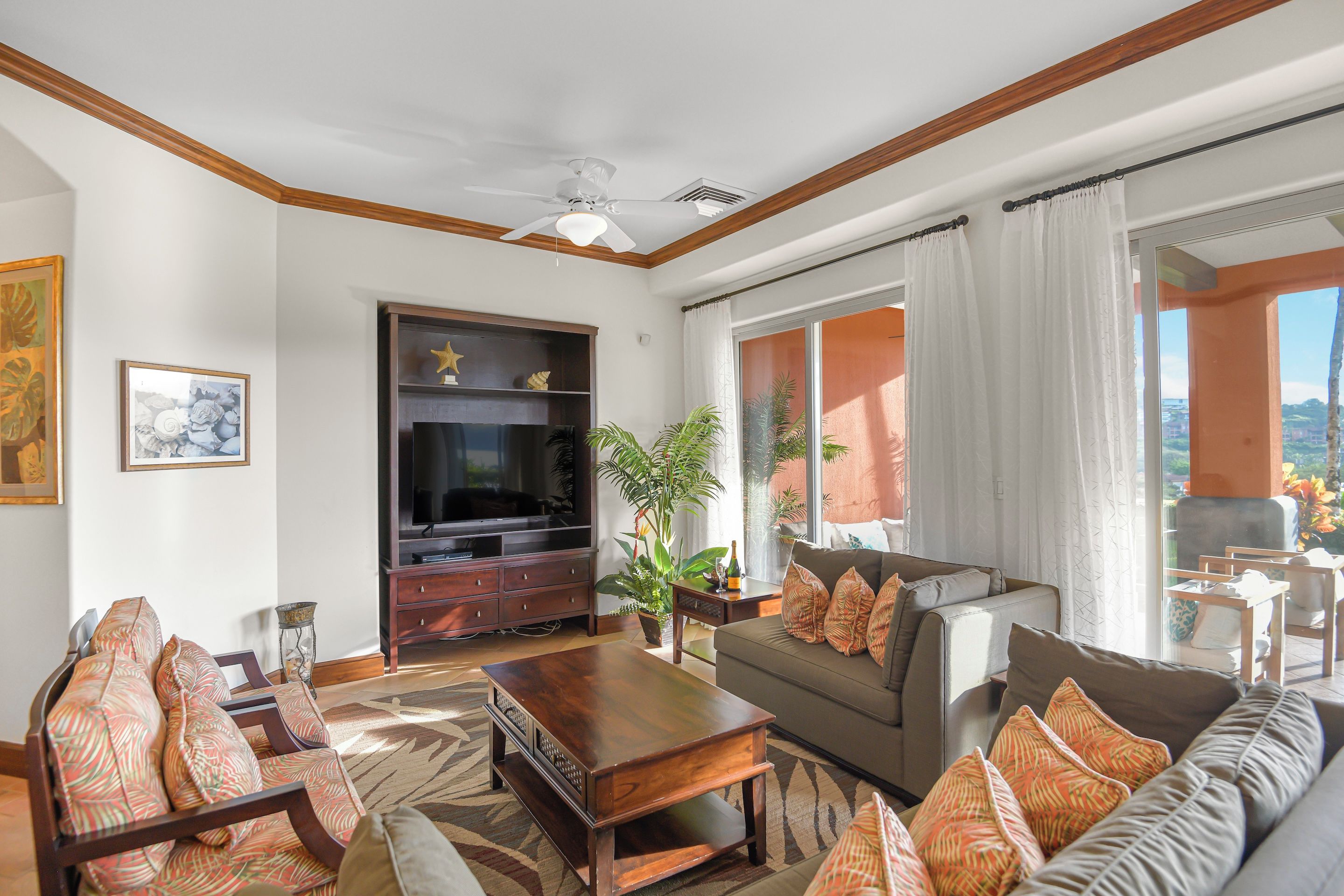 Sala de estar com televisão, sofá, poltronas, ventilador de teto e uma janela grande com cortinas.