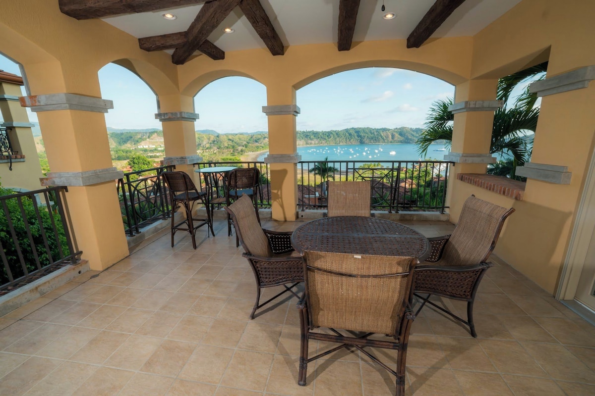 A covered outdoor patio with a round table and chairs, a view of a body of water, and a mountainous landscape in the background.