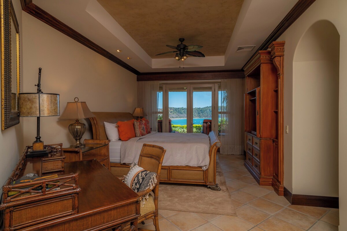 A spacious bedroom with a large bed, a desk, and a chair. There is a view of the outdoors through the windows.