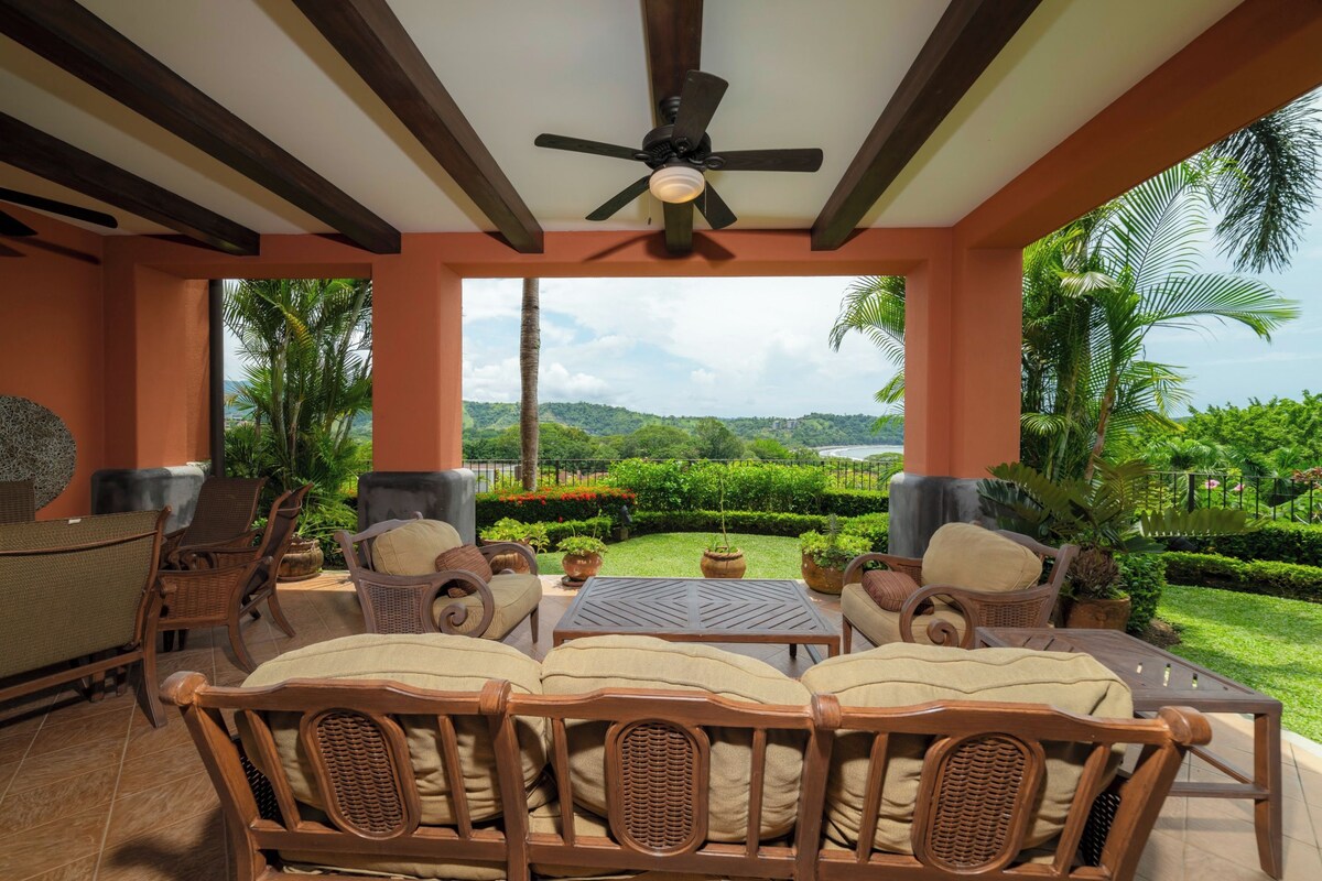 A covered outdoor seating area with wicker furniture, a ceiling fan, and a view of a lush garden and distant hills.