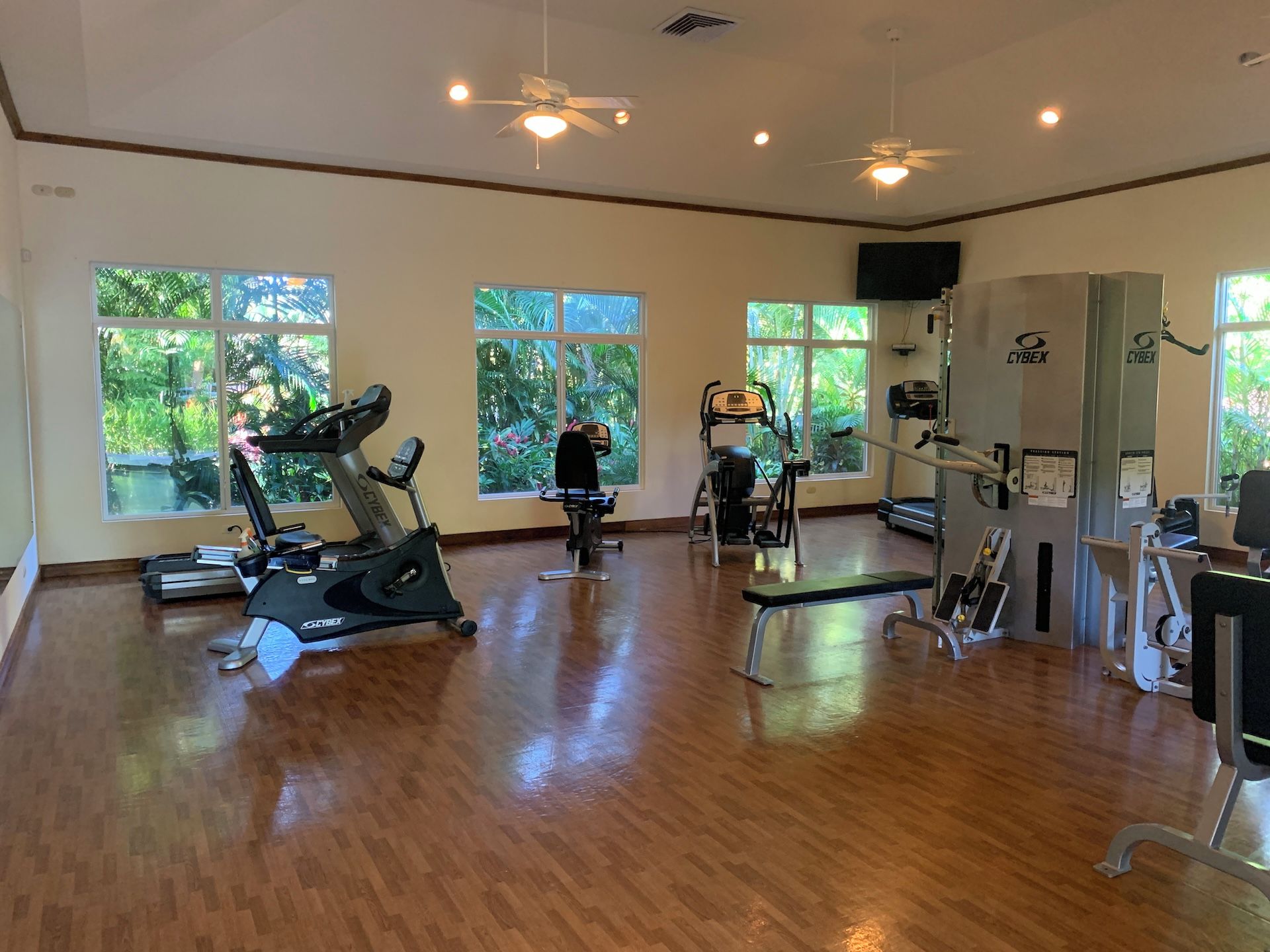 Une salle de sport spacieuse, dotée de divers appareils d’exercice, de grandes fenêtres offrant une vue sur la verdure et de ventilateurs de plafond.